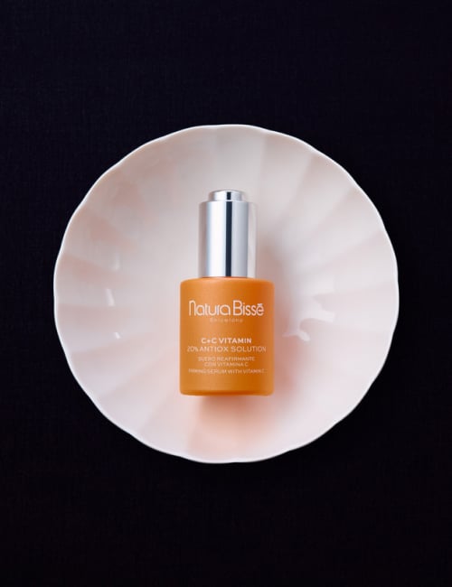 Woman smiling and holding Natura Bissé C+C Vitamin Antioxidant Solution serum, wearing an orange top. Skincare product featured in bestsellers.
