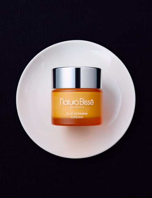 Woman smiling and holding Natura Bissé C+C Vitamin Cream moisturizer.. Skincare product featured in bestsellers.