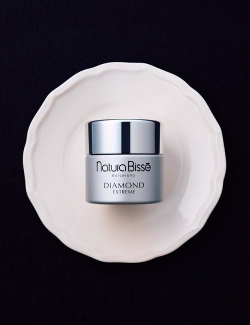 Woman with a slight smile holding Natura Bissé's Diamond Extreme Cream moisturizer with a rich texture. Skincare product featured in bestsellers.
