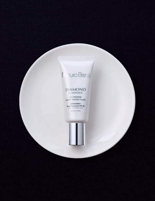 Woman smiling and holding Natura Bissé's Diamond Luminous SPF Sunscreen certified by the Skin Cancer Foundation. Skincare product featured in bestsellers.