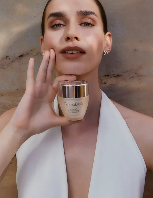 Mujer sonriendo y sosteniendo la crema hidratante Inhibit High Definition Lifting Cream. Producto presentado en nuestros bestsellers.