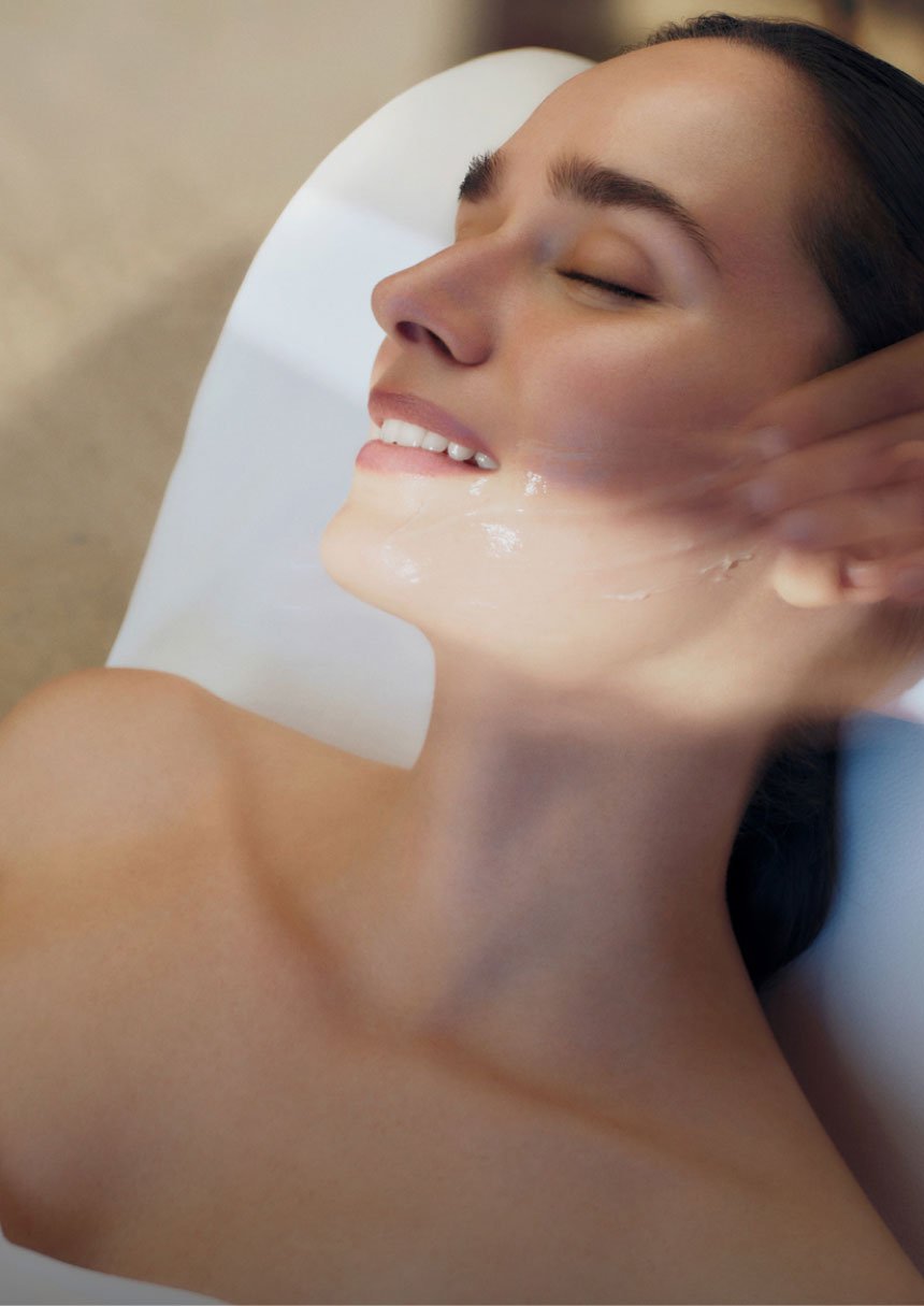Brunette woman smiles while an aestheticien applies moisturizing cream on her face