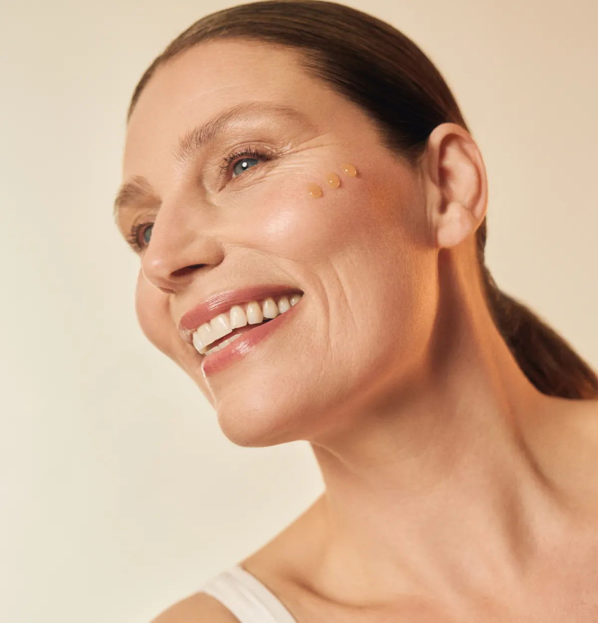 smiling woman in her fifties with a clean girl look: tight ponytail and minimal makeup is applying eye cream